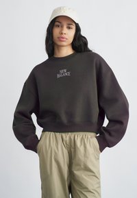 Νεαρή γυναίκα με μακριά σκούρα μαλλιά φοράει μπεζ καπελάκι, σκούρο καφέ φούτερ New Balance και μπεζ παντελόνι, στέκεται με τα χέρια στις τσέπες.