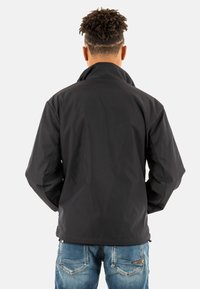 Giacca nera impermeabile con colletto alto, texture morbida e polsini elasticizzati. Indossata con jeans in denim blu, mostra un fit casual.