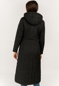 Kobieta w długim czarnym pikowanym płaszczu z kapturem, pokazana od tyłu na jednolitym jasnym tle.