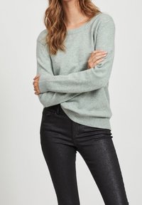 Svetle zelený pletený svetr s kulatým výstřihem a žebrovanými manžetami, v kombinaci s přiléhavými černými džínami s jemným leskem.