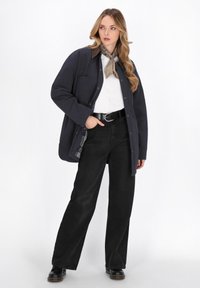 Donna che indossa una giacca nera oversize, una camicia bianca, pantaloni neri a gamba larga, una cintura nera, stivali neri e una sciarpa beige a motivi, in piedi con una mano in tasca.