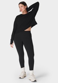 Zwart cropped top met lange mouwen en een losse pasvorm, gecombineerd met zwarte high-waisted leggings. Witte sportvrienden en opgerolde enkelsokken zijn inbegrepen.
