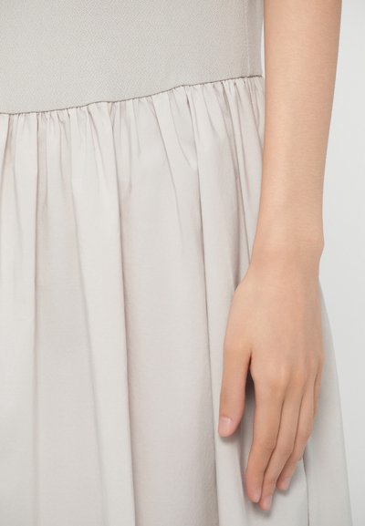 Šviesiai kreminė suknelė su tekstūruotu liemeniu, surinkta sijonu ir lygiu audiniu, o ranka remiasi ant sijono krašto.