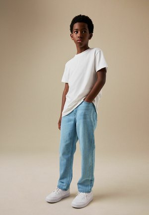 Boy standing with one hand in pocket, wearing a white t-shirt, light blue jeans, and white sneakers against a plain beige background.