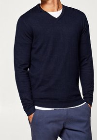 Navy blue v-neck sweater, knit texture, long sleeves, ribbed cuffs and hem, worn over a white t-shirt, paired with light blue trousers.