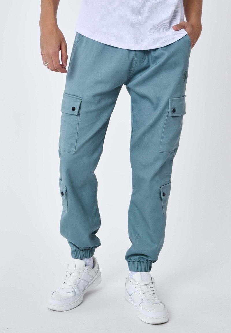 Homme portant un pantalon cargo jogger bleu sarcelle avec des poignets élastiques et des baskets blanches, debout avec une main dans la poche sur un fond uni.