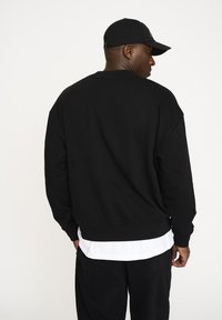 Sort oversized sweatshirt med ribbet kant, rund hals og sænkede skuldre, lagdelt over en hvid skjorte. Ensartet tekstur, ingen mønstre.
