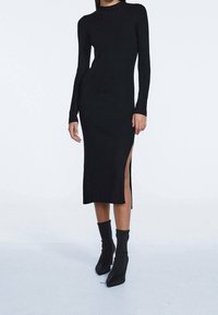 Abito a maniche lunghe nero in maglia a coste, con uno spacco laterale, scollatura alta, design aderente e morbida texture. Abbinato a stivaletti neri alla caviglia.