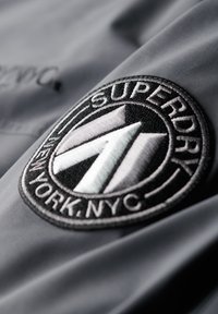 Tissu gris avec un patch circulaire brodé noir portant le texte "SUPERDRY" et un design géométrique en blanc et gris.