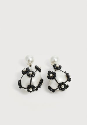 Boucles d'oreilles gouttes de perle blanche décorées de grappes de fleurs en perles noires et de petites perles blanches, sur un fond clair uni.