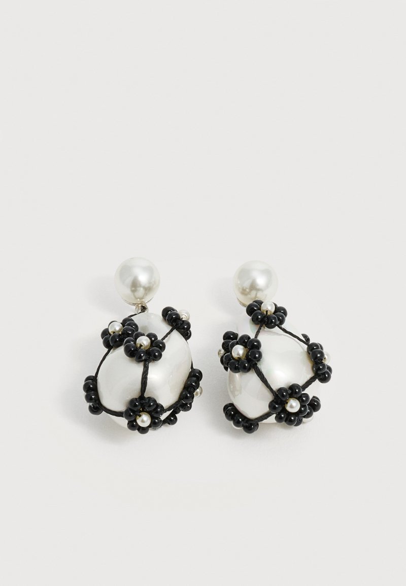 Boucles d'oreilles gouttes de perle blanche décorées de grappes de fleurs en perles noires et de petites perles blanches, sur un fond clair uni.