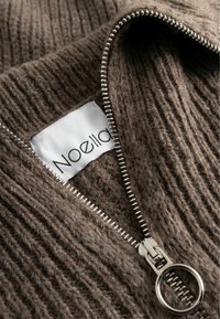 Brūns teksturēts adījums ar sudraba rāvējslēdzēju un apļa vilkšanas elementu. Iekļauj baltu birku ar "Noella" logo.