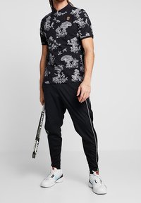 Zwarte poloshirt met een wit bloemenpatroon, korte mouwen, gecombineerd met zwarte, taps toelopende broek met een zijstreep. Witte sportschoenen, terwijl er een stok wordt vastgehouden.