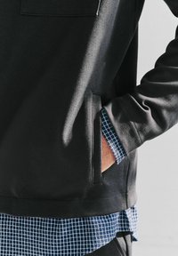 Zwart sweatshirt met zichtbare zak, textuurstof en een marineblauw geruit patroon dat onder de rand tevoorschijn komt. Geschikt voor casual gebruik.