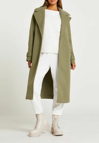 Cappotto lungo verde oliva in tessuto testurizzato, abbinato a un top bianco e pantaloni bianchi. Completato da stivali chunk beige con lacci.