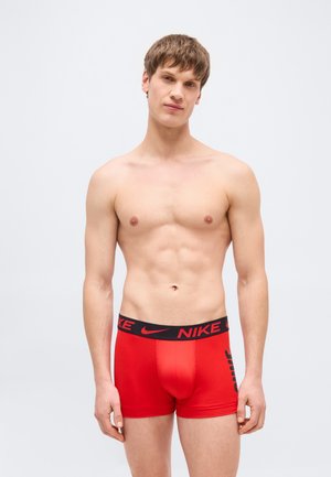 Jeune homme torse nu aux cheveux courts bruns portant un boxer rouge Nike, debout et faisant face, sur un fond clair uni.