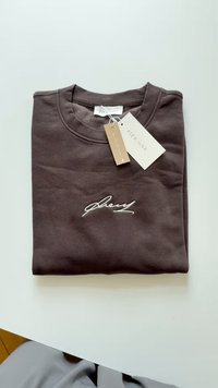 Brown folded sweatshirt made of cotton, featuring a round neckline, embroidered signature detail in white, and hang tags attached.