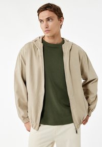 Chaqueta de cremallera beige con capucha, puños elásticos y un ligero brillo, llevada sobre una camiseta verde oscuro. Tela suave, corte casual.