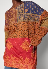 Top de manga larga con un diseño de patchwork que incluye estampados animales en marrón y naranja, patrones florales en rojo, y acentos en azul y dorado.