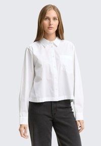 Camicia bianca a maniche lunghe, cropped, con colletto classico e una tasca sul petto. Presenta una sottile texture e un orlo dritto.