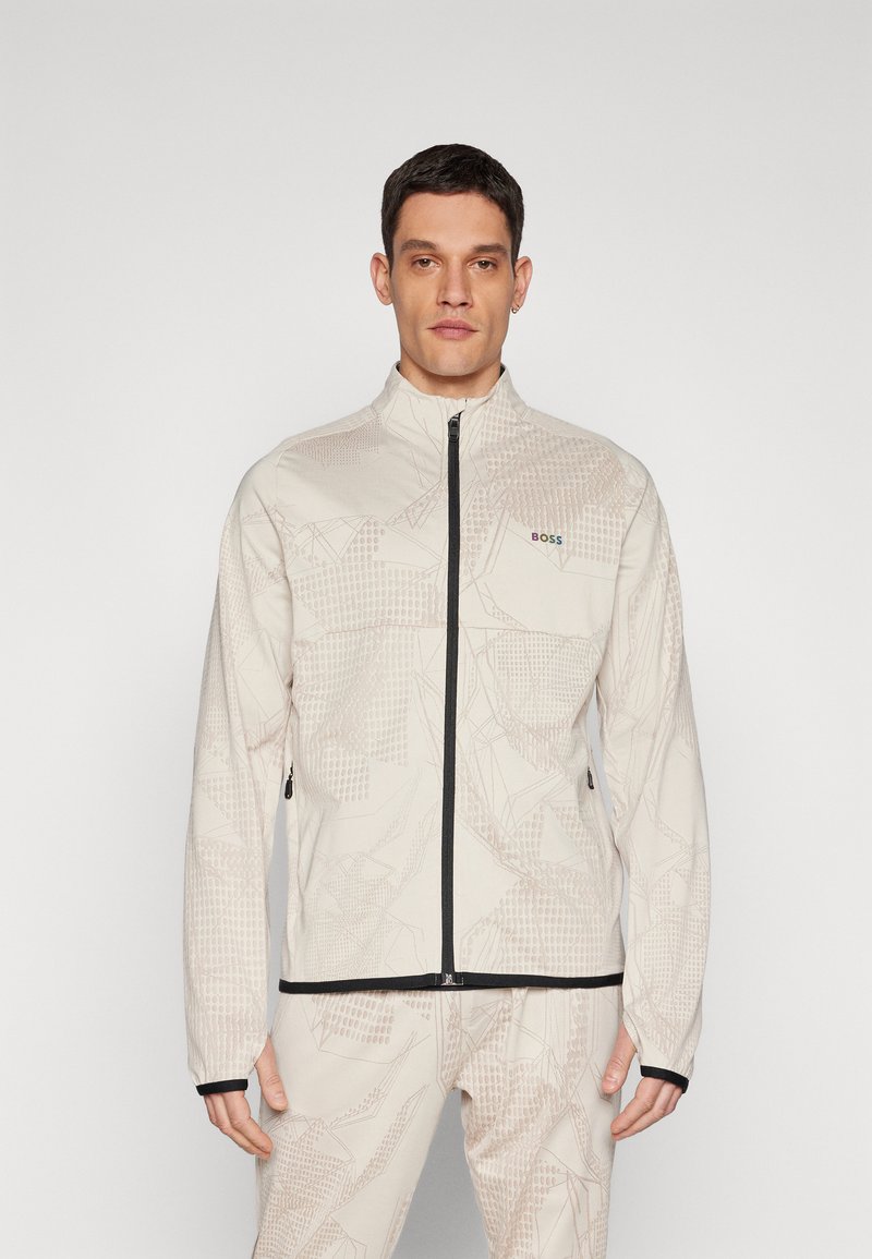Lys beige zip-up jakke med geometriske mønstre, sorte detaljer ved manchetter og bund, lavet af blødt materiale, med høj krave og logo-detalje.
