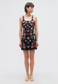 Kvinde med kort mørkt hår, stående barfodet iført en ærmeløs sort kjole med farverig blomsterbroderi, kigger mod højre.