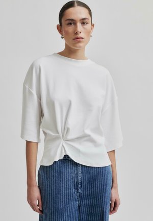 Vit kortärmad topp med snurrad frontdetalj, tillverkad av mjukt tyg, kombinerad med blå vertikalt randiga denimbyxor.