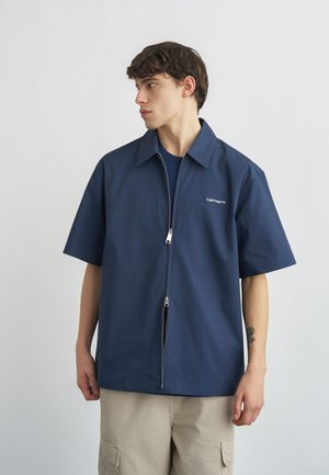 Νέος άνδρας που φοράει μπλε σκούρο κοντομάνικο πουκάμισο με φερμουάρ και μπεζ παντελόνι, κοιτάζοντας προς τα αριστερά, μπροστά σε απλό ανοιχτό φόντο.