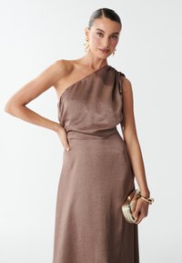Abito satinato marrone con un design a spalla unica, caratterizzato da un dettaglio a nodo. Il modello tiene una borsa color oro con una chiusura.