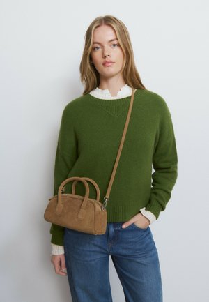 Woman wearing green sweater and white shirt, carrying a small brown crossbody bag, standing against a plain light background.