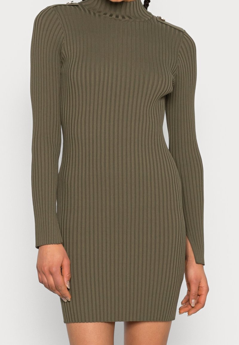 Ribbad långärmad klänning i olivgrön med hög halsringning och sidofickor, med knappdekorationer på ena axeln för detalj och textur.