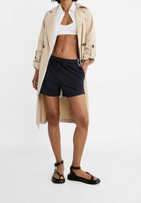 Beige trenčkot, bílý zkrácený top, námořnické šortky na šňůrku a černé sandály. Hladké textury, ležérní střih a vrstvený design.