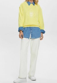 Žlutá mikina s potiskem "100% Organická bavlna", vrstvená přes džínovou košili s knoflíky, doplněná o bílé široké kalhoty a bílé tenisky.