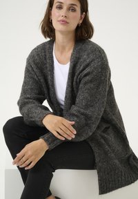 Cardigan moale gri, cu o croială largă, tricot texturat, deschis în față și cu manșete nervurate, purtat peste o cămașă albă și asortat cu blugi negri.