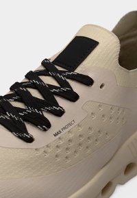 Baskets beige avec une tige texturée, des lacets noirs à rayures blanches et un patch d'accent noir. Comprend un détail de logo sur le côté.