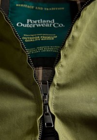 Close-up of a partially zipped olive green jacket revealing a dark green label with white and yellow text inside.
