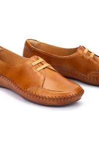 Zapatos de cuero marrón claro con un perfil bajo, que presentan costuras a lo largo de la suela y dos bandas elásticas decorativas en la parte superior. Textura suave.