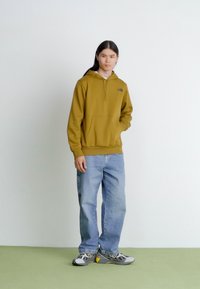 Olive green hooded sweatshirt with a front pocket, paired with light blue jeans and gray sneakers, set against a neutral background.