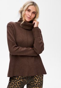 Blondynka w brązowym swetrze z wysokim kołnierzem i spodniach w panterkę, dotykająca kołnierza i patrząca w kamerę.