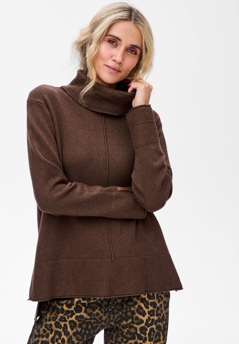 Blondynka w brązowym swetrze z wysokim kołnierzem i spodniach w panterkę, dotykająca kołnierza i patrząca w kamerę.