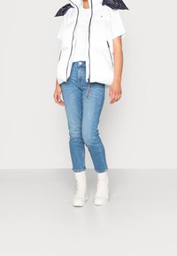 Puchowa, biała kamizelka z granatowymi akcentami, w zestawieniu z jasnoniebieskimi dżinsami i białymi botkami za kostkę. Kamizelka ma wysoki kołnierz i zamek błyskawiczny z przodu.