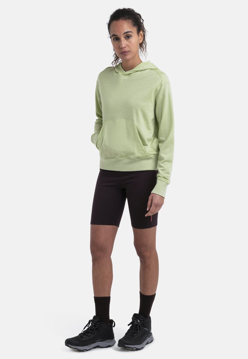 Hoodie verde claro con un bolsillo frontal, combinado con pantalones cortos de ciclismo marrón oscuro. Botas de senderismo negras con suelas texturizadas y calcetines oscuros.