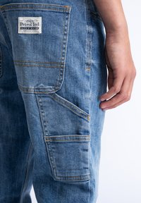 Pantalones cargo de mezclilla azul claro con bolsillos laterales y traseros, que cuentan con costuras amarillas y una etiqueta en el bolsillo trasero.