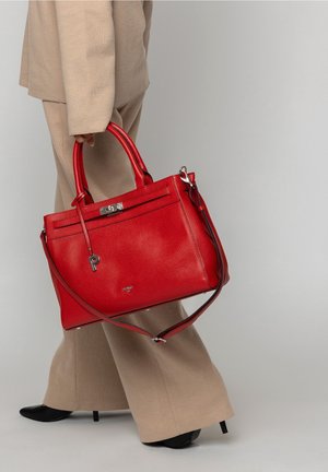 Osoba v béžovém oblečení držící velkou červenou koženou kabelku s horními rukojetěmi a ramenním popruhem, stojící před jednobarevným pozadím.