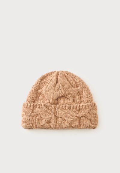 Bonnet en laine beige tricoté avec un revers plié et un motif torsadé, posé sur un fond clair uni.