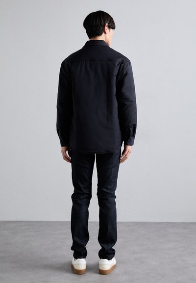 Camicia nera con colletto classico e maniche lunghe, con polsini abbottonati, abbinata a jeans scuri slim-fit e sneakers bianche.