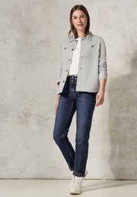 Chaqueta abotonada gris claro con cuello, combinada con jeans oscuros ajustados y zapatillas blancas. Tejido suave con elementos de diseño casual.