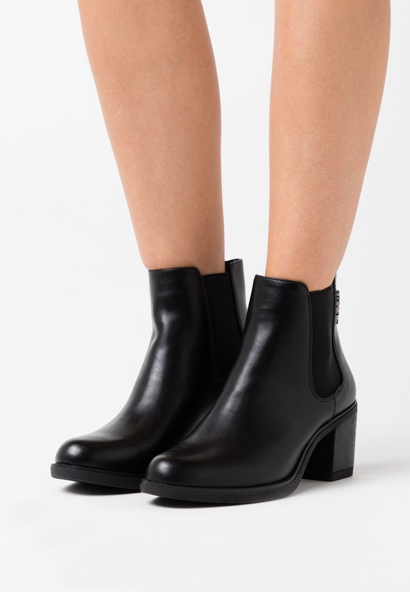 Bottes chevilles en cuir noir avec panneaux latéraux en élasthanne et talons carrés portées sur des jambes nues sur fond blanc.