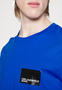 Zila kokvilnas T-kreklu ar īsām piedurknēm, ar spīdīgu melnu logotipa uzlīmi, kurā rakstīts "KARL LAGERFELD JEANS." Gluda tekstūra, klasiskā piegriezuma.
