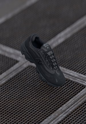 Musta New Balance 1600 -tennari roikkuen teksturoidun metalliritilälattian yläpuolella, jonka vinot viivat muodostavat ruudukkomallin.